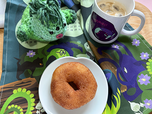 flat lay of Brewing Up the Magic 2026 tea towel with a mood mug, a donut, and a green, crochet monster.